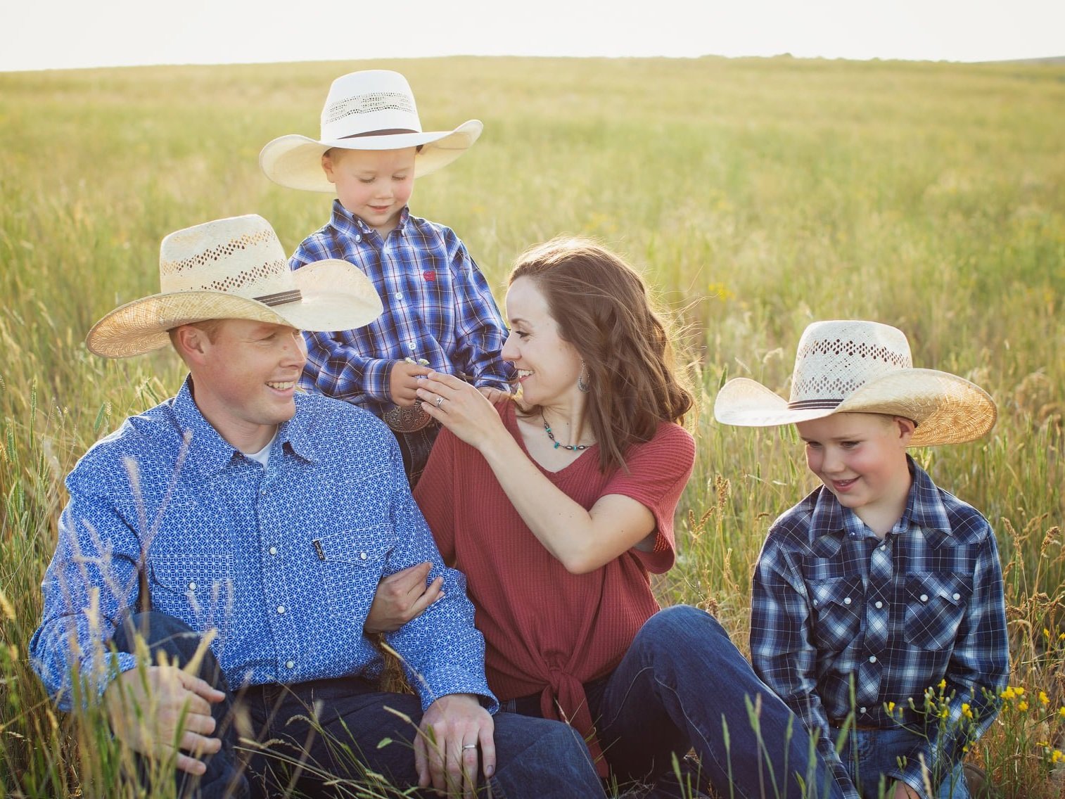 A happy family of four sitting in a grassy field during sunset, wearing cowboy hats and casual western attire. The father and mother are interacting with their two young sons, who are also dressed in plaid shirts, creating a warm and joyful outdoor family moment.