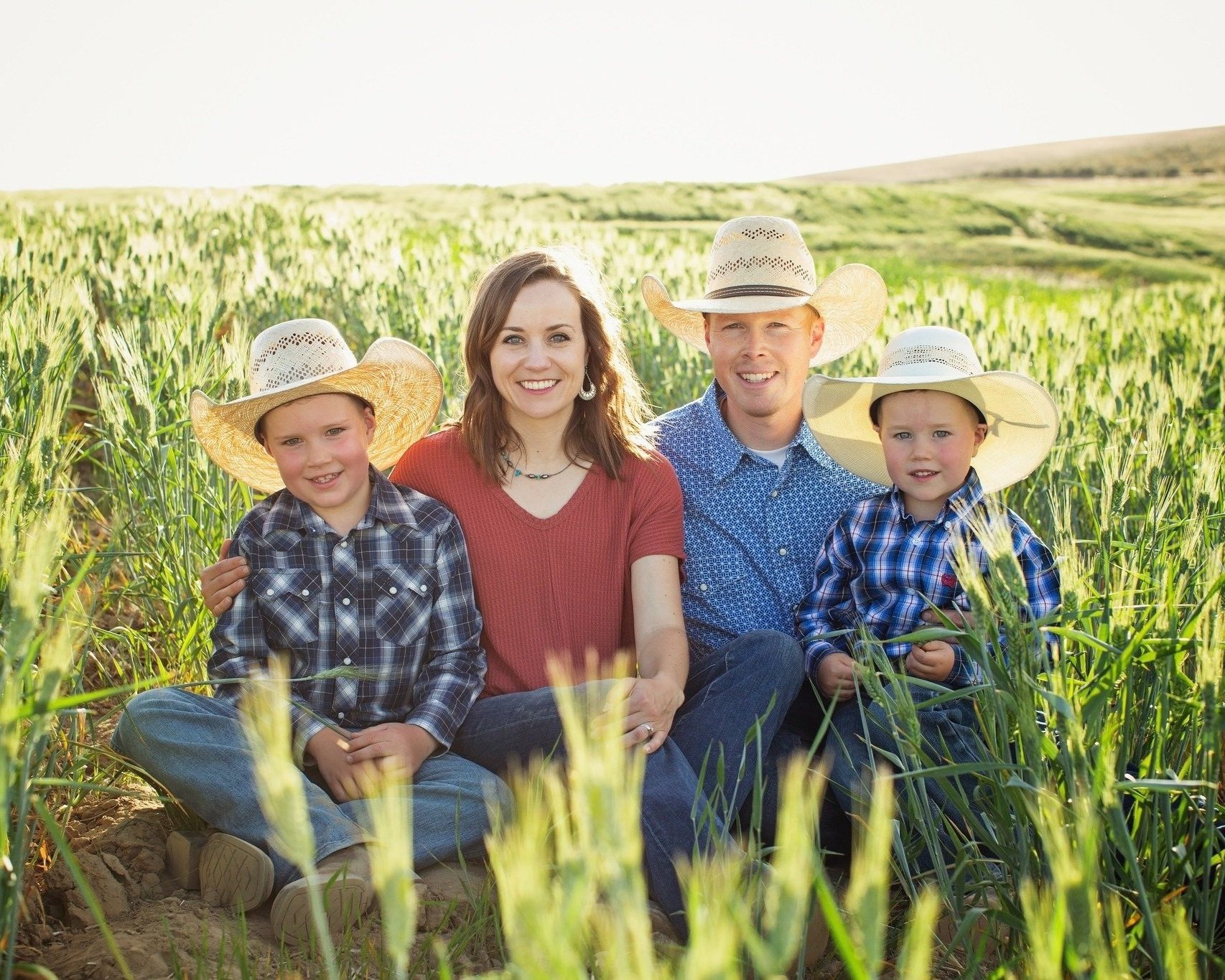 Family portrait in a green wheat field, featuring a woman and three children wearing straw cowboy hats. The scene captures a warm, sunny day, highlighting a joyful family moment amidst nature.