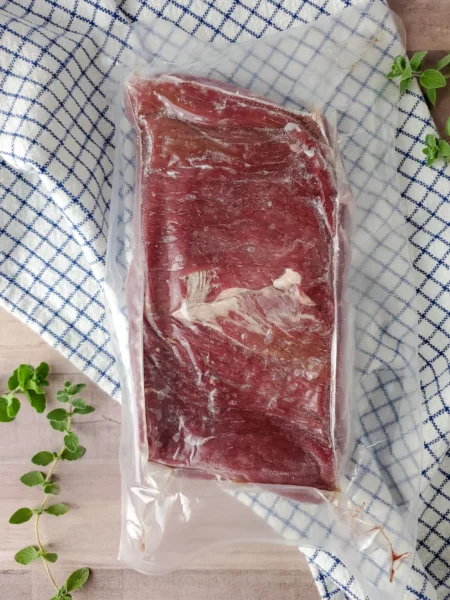 Vacuum-sealed slab of beef resting on a blue and white checkered cloth, surrounded by fresh oregano leaves, ideal for meal preparation or cooking.