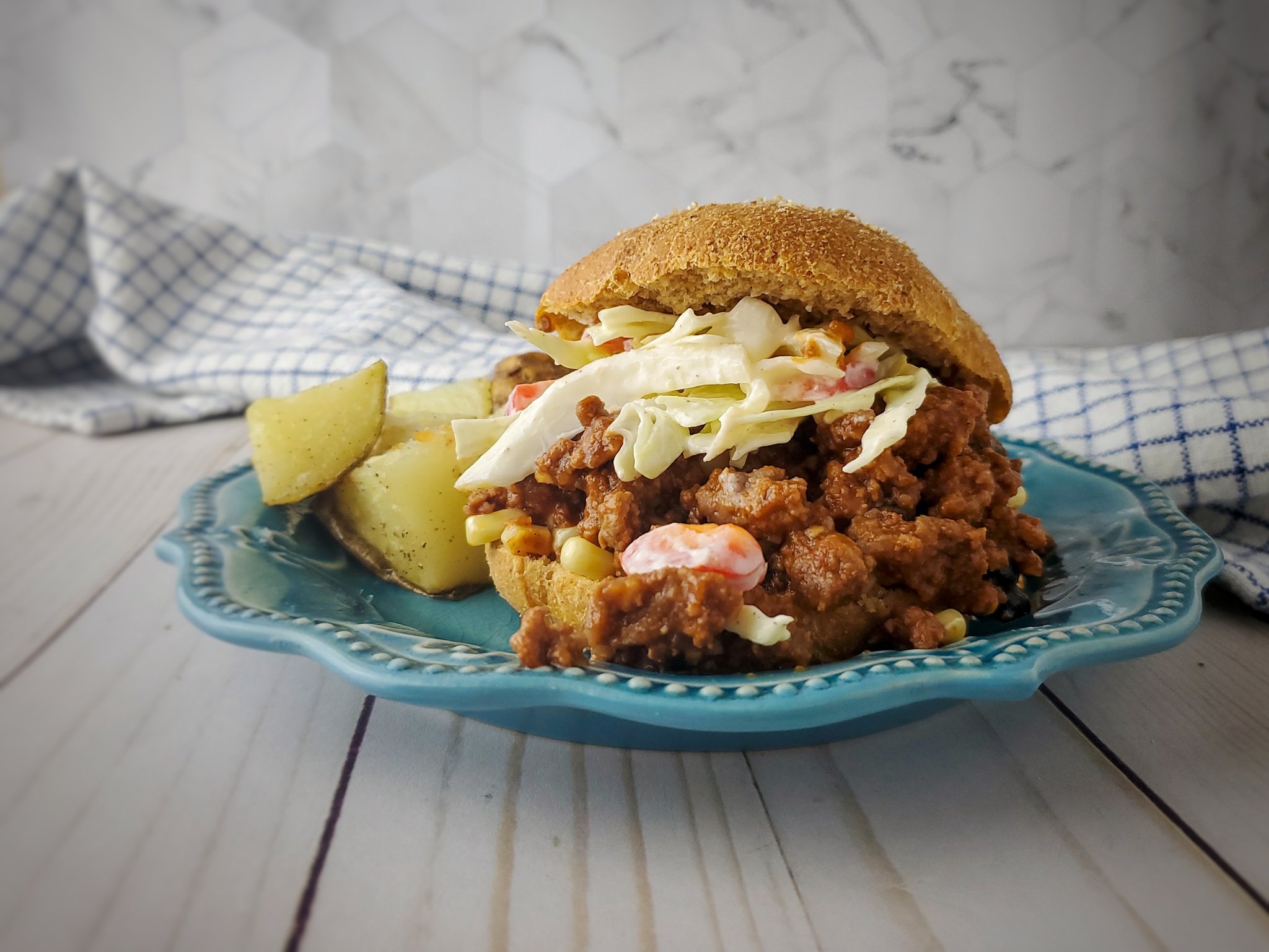 Sloppy joe sandwich topped with coleslaw served on a blue plate alongside seasoned potato wedges, with a checkered napkin in the background.